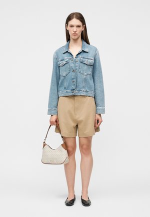 Γυναίκα που φοράει γαλάζιο τζιν μπουφάν και μπεζ σορτς, κρατώντας μια λευκή και καφέ τσάντα, στέκεται μπροστά σε ένα απλό φόντο.