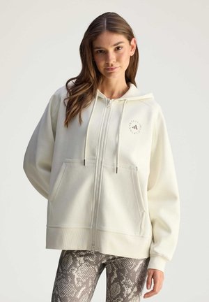 White zip-up hoodie with a front logo, large pockets, and drawstring hood, paired with grey snakeskin patterned leggings.