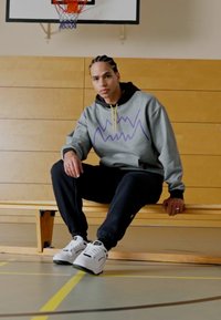 Sudadera gris con gráfico púrpura, pantalones negros y zapatillas blancas, sentado en un banco de madera en un gimnasio con una canasta de baloncesto al fondo.
