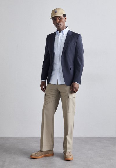 Blazer marine sur une chemise à boutons bleu clair, pantalon cargo beige avec poches latérales et chaussures beiges. Une casquette beige avec un logo complète le look.