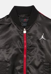 Casaco bomber em satin preto com um fecho vermelho, apresentando um logo branco da Jordan no lado esquerdo do peito e colarinho canelado preto. Acentos de tecido texturizado.