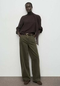 Top de manga larga marrón oscuro con cuello alto y detalle anudado, acompañado de pantalones de pierna ancha en verde oliva. Cinturón marrón y calzado suave.