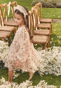 Laps heleroosas lillelises kleidis kuldsete detailidega, kõndimas puidust toolide rea lähedal ja ümbritsetud valgetest õitest rohelisel murul.