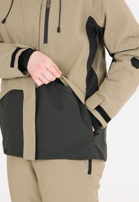 Giacca da esterno beige e nera con materiale impermeabile, tasca con zip, polsini regolabili e un design elegante e aderente.