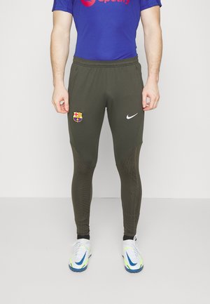 Pantalones deportivos Nike en verde oliva oscuro. Presentan un logo de Barcelona, paneles texturizados y un diseño de ajuste slim; combinados con zapatos blancos con detalles en neón.