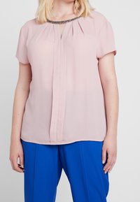 Blus i mjuk rosa chiffon med kort ärm, plisserad fram, nyckelhålsurringning och dekorerad krage; matchad med klara blå byxor.