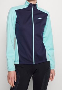 Chaqueta de cremallera completa en azul marino y azul claro con cuello alzado, hecha de una tela ligera. Presenta un logo en el pecho y mangas en contraste.