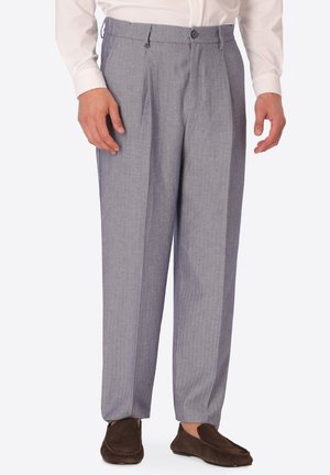 Uomo con pantaloni da abito plissettati grigio chiaro, camicia bianca e mocassini marrone scuro, in piedi davanti a uno sfondo semplice.