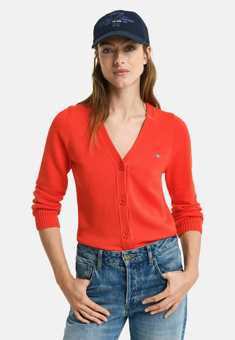 Red knit cardigan with a v-neck and five buttons, paired with blue denim jeans. The model also wears a navy cap with white text.