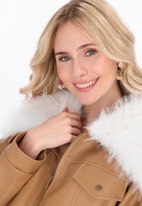 Giovane donna con capelli biondi sorride, indossando una giacca in pelle beige con colletto in pelliccia bianca e orecchini a cerchio dorati con ciondoli pendenti.