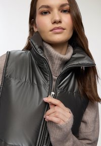 Gilet imbottito grigio scuro trapuntato con colletto alto e chiusura con zip, indossato sopra un maglione a collo alto lavorato a maglia di un beige chiaro.