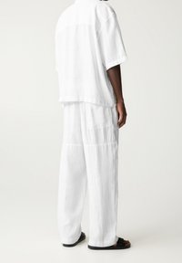 Personne debout portant un pantalon ample en lin blanc et une chemise à manches courtes assortie, associée à des sandales noires, sur un fond blanc.