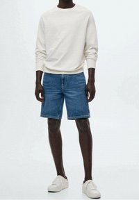 Tenue décontractée avec une chemise à manches longues crème et un short en denim bleu. Des baskets blanches complètent le look. Style minimaliste avec une coupe confortable.