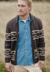 Mørkebrunt strikket cardigan med beige mønstre, featuring en sjalskrave og lynlåsdetaill, båret over en lys blå chambray skjorte.