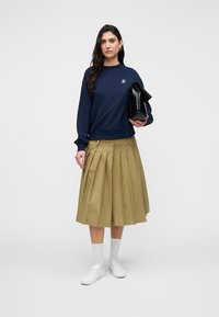 Μπλε φούτερ με στρογγυλή λαιμόκοψη και λογότυπο, συνδυασμένο με χακί πλισέ φούστα. Άσπρα αθλητικά παπούτσια και λευκές κάλτσες μέχρι τον αστράγαλο ολοκληρώνουν την εμφάνιση.