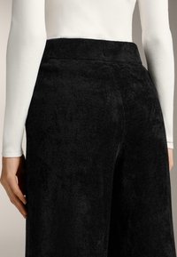 Čierne teplákové nohavice z froté tkaniny s rebrovaným pásom. Textúra vyzerá mäkko a nadýchane, čo zaisťuje uvoľnený strih. Bočný pohľad ukazuje hladké kontúry.