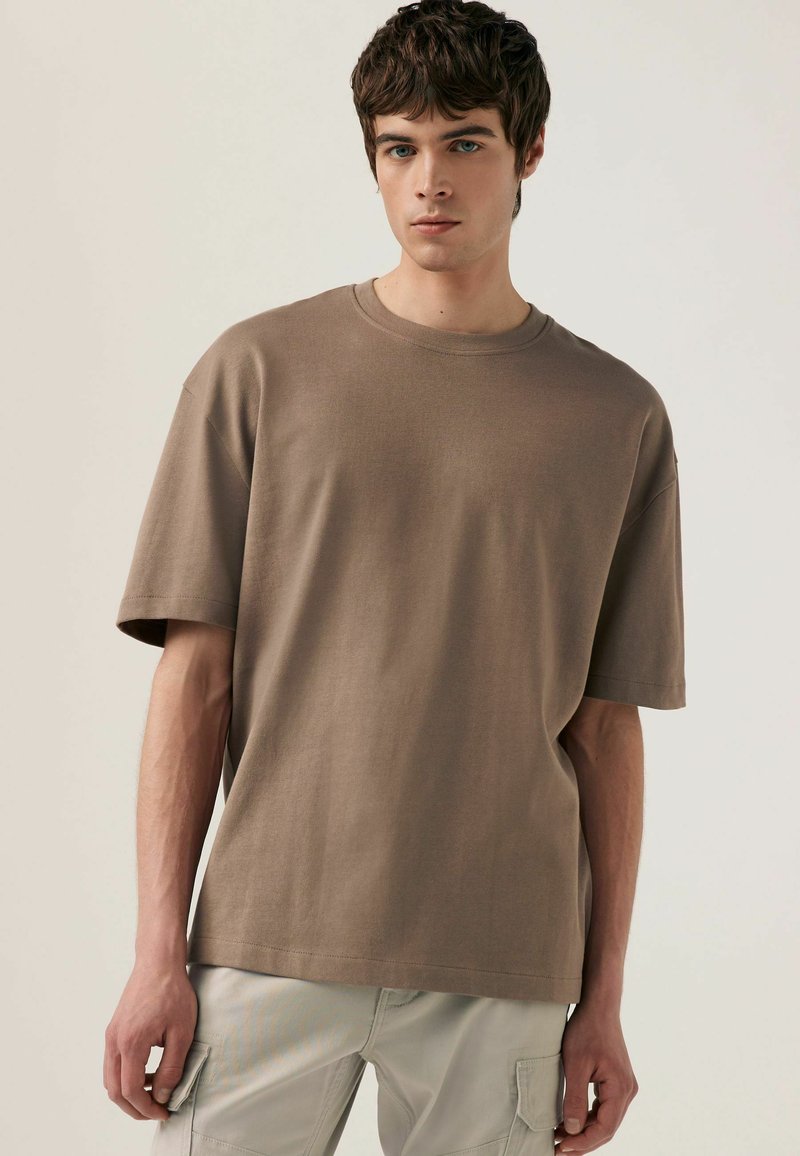 Tricou scurt cu mânecă scurtă, de culoare maronie, cu guler rotund. Fabricat dintr-un material moale și texturat; are o croială lejeră și detalii de cusătură vizibile.