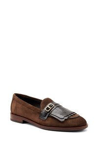 Mocassino in suede marrone con un accento e fibbia in pelle nera, caratterizzato da un dettaglio frangiato a scallop. Suola piatta e design a punta arrotondata.