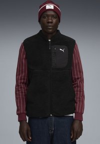 Schwarze Fleece-Weste mit einem Reißverschluss vorne und einer Brusttasche, kombiniert mit einem bordeauxrot gestreiften Langarmshirt und einer passenden bordeauxrot Mütze.