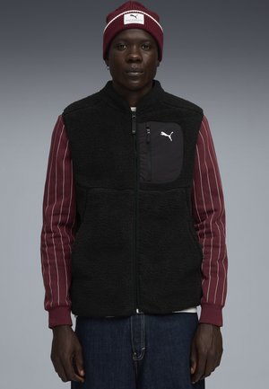 Schwarze Fleece-Weste mit einem Reißverschluss vorne und einer Brusttasche, kombiniert mit einem bordeauxrot gestreiften Langarmshirt und einer passenden bordeauxrot Mütze.