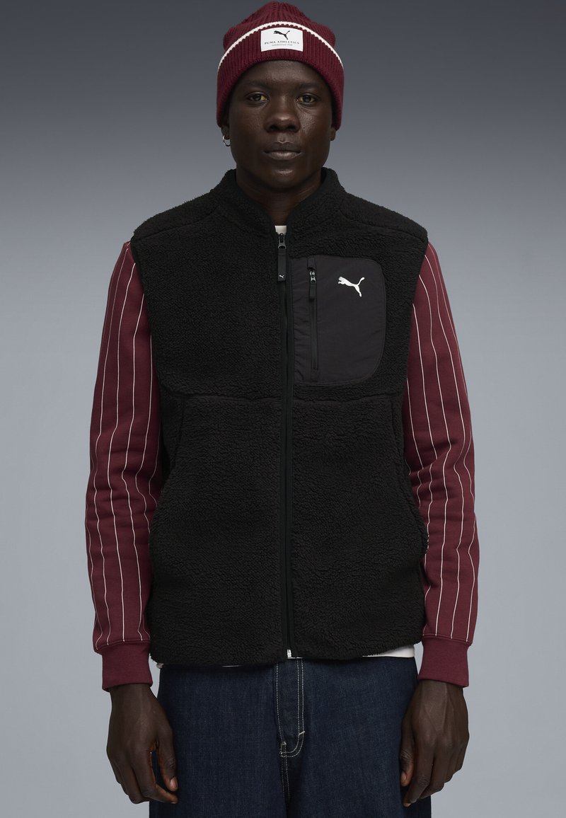 Schwarze Fleece-Weste mit einem Reißverschluss vorne und einer Brusttasche, kombiniert mit einem bordeauxrot gestreiften Langarmshirt und einer passenden bordeauxrot Mütze.