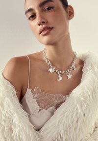 Woman wearing a silver charm necklace with moon and star pendants, a lace-trimmed camisole, and a white fluffy coat.