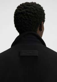 Cappotto di lana nera con colletto alto, con un'etichetta rettangolare che riporta la scritta "HUGO". La texture è morbida, con una finitura liscia e lucida.