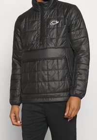 Zwarte gewatteerde pufferjack met hoge kraag, halve rits en voorkantzak. Bevat een wit Nike-logo op de borst. Soepele textuur.