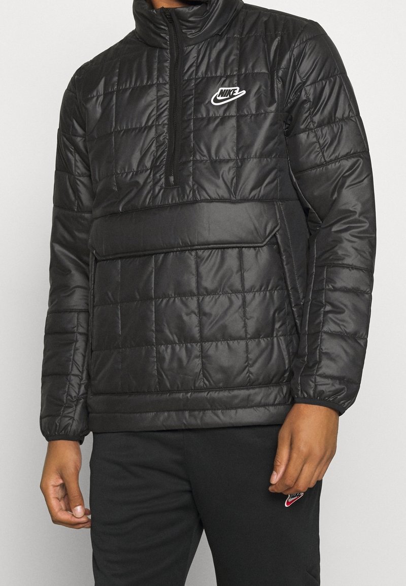 Zwarte gewatteerde pufferjack met hoge kraag, halve rits en voorkantzak. Bevat een wit Nike-logo op de borst. Soepele textuur.