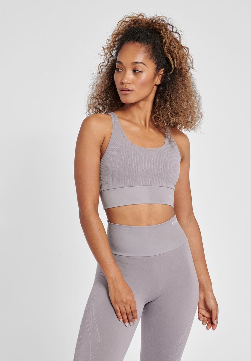 Kvinde med krøllet hår iført matchende lilla sports-bh og høj-taljede leggings, stående mod en ensfarvet lys baggrund, kigger mod venstre.