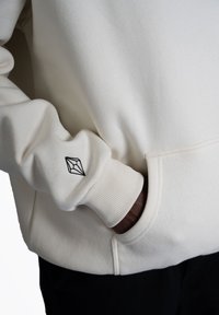 Hoodie beige chiaro realizzato in tessuto morbido e testurizzato. Presenta polsini a coste e un piccolo logo nero ricamato sulla manica destra.