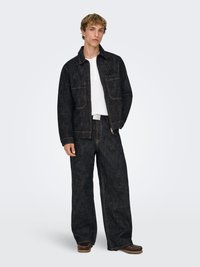 Donker denimjack met twee voorzakken, gecombineerd met donkere jeans met wijde pijpen. Witte henley shirt eronder. Contrasterende stikseldetails zichtbaar.