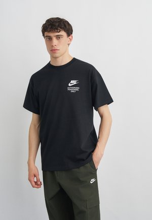 Mladić kovrčave tamne kose u crnoj Nike majici s natpisom "Authorized Personnel Only" i tamnozelenim Nike cargo hlačama.