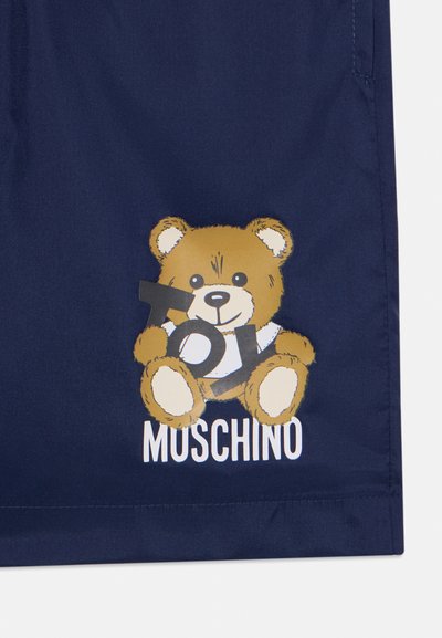 Jūras zila auduma šorti ar karikatūras lācīti, uz kura redzami vārdi "Toy" un "Moschino" baltā krāsā, piedāvājot rotaļīgu un krāsainu dizainu.