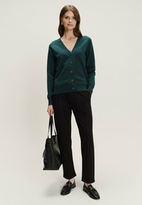 Grüner Cardigan mit V-Ausschnitt und Knopfverschluss, kombiniert mit schwarzen Hosen und Slippern. Modell hält eine schwarze Tote-Tasche. Glatte Textur.