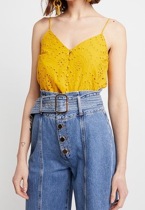 Žena má na sobě žlutý bezrukávový top s průstřihy a knoflíky a modré džíny s vysokým pasem, širokým páskem a velkou přezkou.