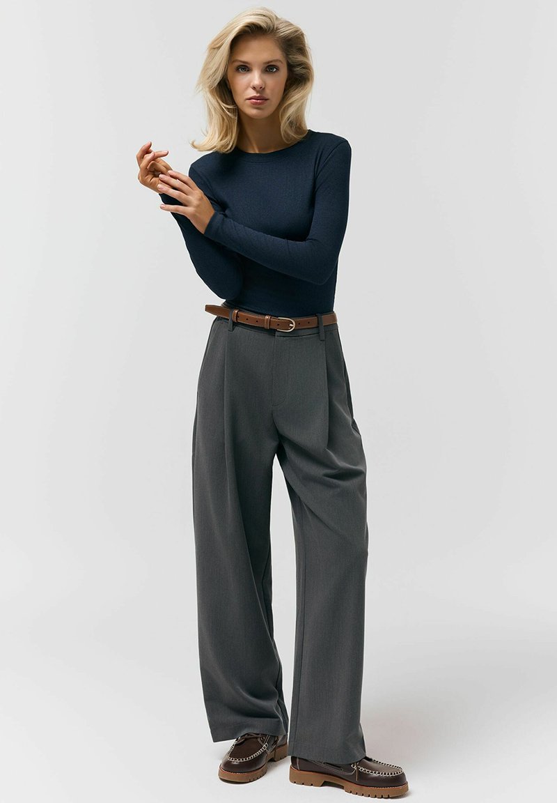 Kobieta o blond włosach, ubrana w granatową bluzkę z długim rękawem, szare szerokie spodnie z brązowym paskiem oraz brązowe mokasyny, stojąca na jednolitym tle.