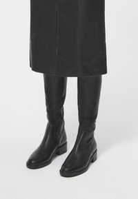 Botas de cuero lisas hasta la rodilla en color negro, con tacón cuadrado y paneles laterales elásticos para facilitar el uso. Diseño simple sin adornos.