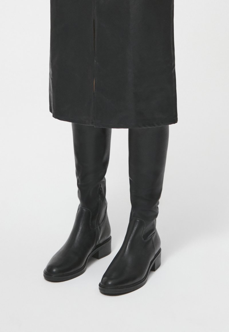 Botas de cuero lisas hasta la rodilla en color negro, con tacón cuadrado y paneles laterales elásticos para facilitar el uso. Diseño simple sin adornos.