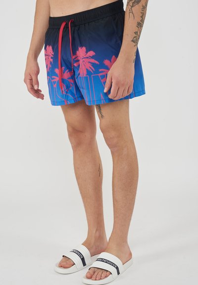 Male wearing blue and black gradient swim shorts with pink palm tree print, red drawstring, and white Armani slides standing on white floor.
