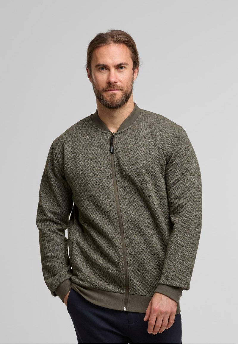 Oliven grøn bomberjakke med et tekstureret strikkemønster, lynlås foran, ribbede manchetter og kant. Stylet med mørke bukser.