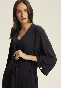 Czarna, prążkowana sukienka o kroju kopertowym z dekoltem w kształcie litery V, szerokimi rękawami i związanym w talii paskiem. Zawiera małą metkę na rękawie dla detalu.