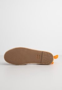 Semelle d'une chaussure marron TOMS avec une semelle en caoutchouc texturée et un talon duveteux, affichant le logo de la marque et des pampilles orange à l'arrière.