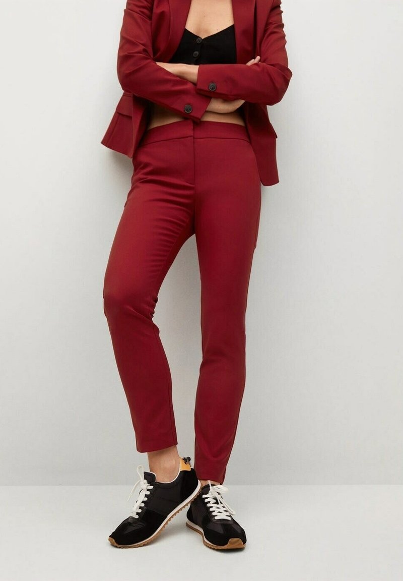 Personne portant un costume rouge ajusté avec une veste boutonnée et un pantalon fuselé, assorti de baskets noir et blanc, debout les bras croisés.