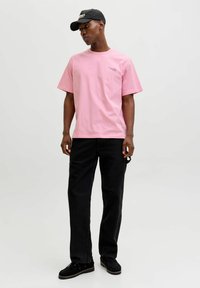 Rosa Baumwoll-T-Shirt mit kurzen Ärmeln und kleinem Logo. Kombiniert mit schwarzen Jeans und schwarzem Schuhwerk. Insgesamt schlichtes Design.