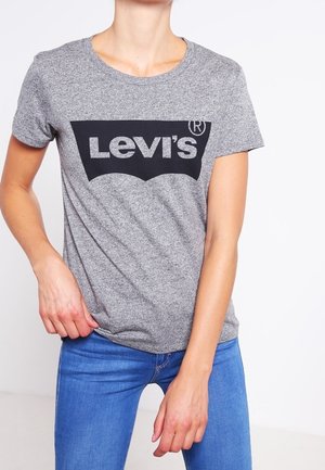 Osoba nosząca szarą koszulkę Levi's i niebieskie dżinsy, stojąca z jedną ręką trzymającą brzeg koszulki.