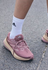 Lichtroze sportschoen met een gestructureerde mesh bovenkant, beige zool en grijze accenten. Draag met witte crew-sokken met zwart logo.