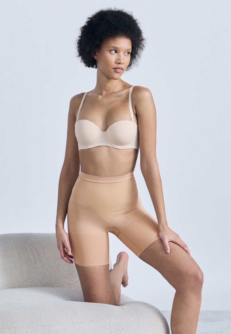 Γυναίκα με σγουρά μαλλιά που φοράει στράπλες μπεζ σουτιέν και μπεζ ψηλόμεσα σορτς σμίλευσης, γονατισμένη σε μια κυρτή ανοιχτό γκρι καναπέ.