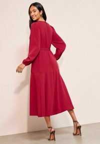 Rochie roșie cu mâneci lungi, cu umeri r gathered, talie ajustată și fustă fluidă. Dispune de un decolteu în fund cu închidere prin buton. Asortată cu sandale cu toc înalt de culoare maronie.