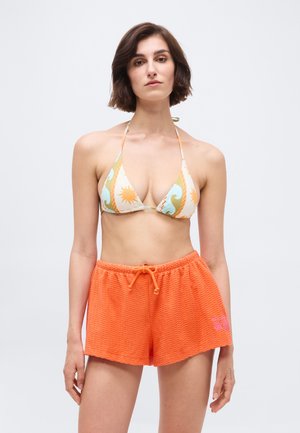 Femme portant un haut de bikini à motifs avec des dessins de soleil et de vagues, ainsi qu'un short orange vif texturé à cordon de serrage, sur un fond uni.
